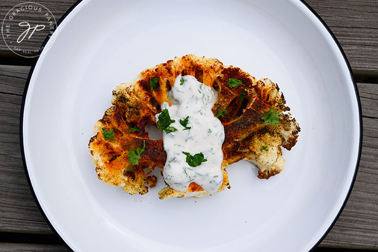 A single Air Fryer Cauliflower Steak sits on a white plate with yogurt sauce spooned over the top.