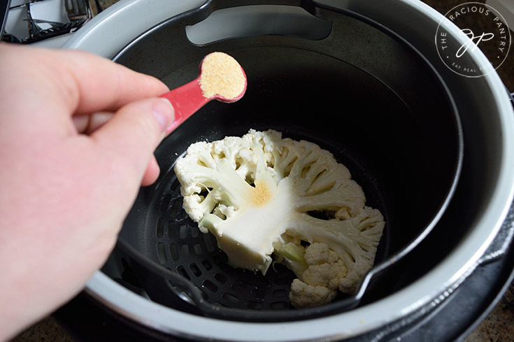 Sprinkling garlic powder over a raw cauliflower steak sitting in an air fryer.