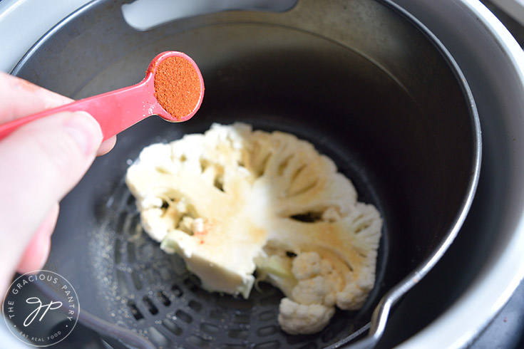 Sprinkling smoked paprika over a raw cauliflower steak sitting in an air fryer.