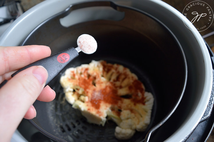 Sprinkling salt over a raw cauliflower steak sitting in an air fryer.