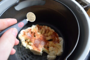 Sprinkling pepper over a raw cauliflower steak sitting in an air fryer.