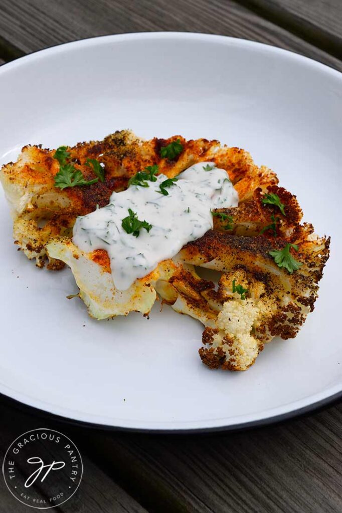 A single Air Fryer Cauliflower Steak on a white plate garnished with yogurt sauce and some fresh parsley.