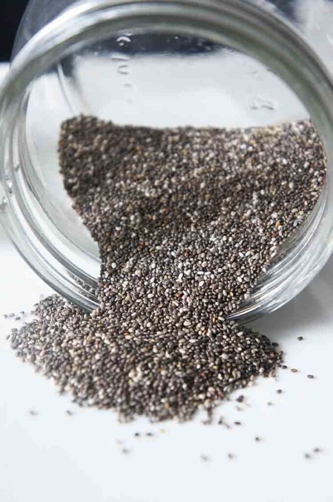 A glass jar of chia seeds lays tipped over on a white surface.