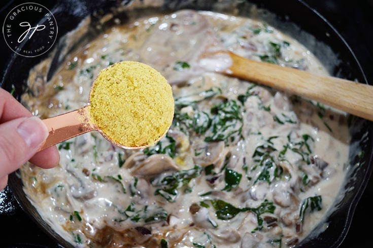 Adding nutritional yeast to Creamy Kale And Mushroom Pasta sauce in a skillet.