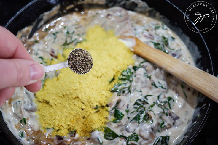 Adding ground pepper to Creamy Kale And Mushroom Pasta sauce in a skillet.