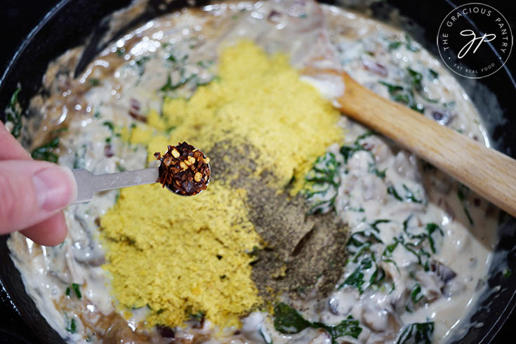 Adding crushed red pepper flakes to Creamy Kale And Mushroom Pasta sauce in a skillet.
