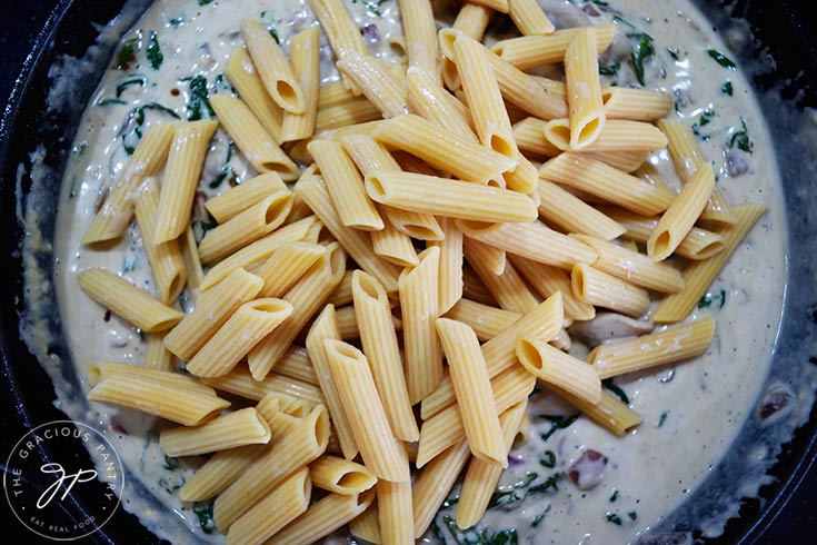 Adding cooked penne pasta to Creamy Kale And Mushroom Pasta sauce in a skillet.