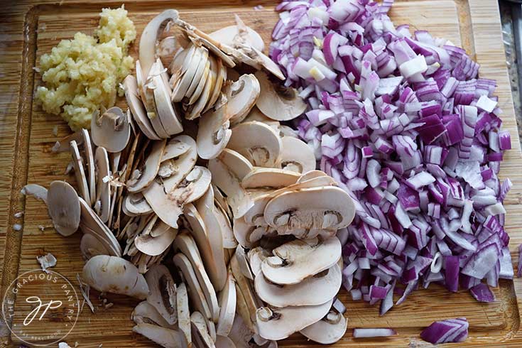 Chopped, red onions, sliced mushrooms and pressed garlic piled up on a wood cutting board.