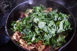 Chopped kale added to onions and mushrooms in a cast iron skillet.