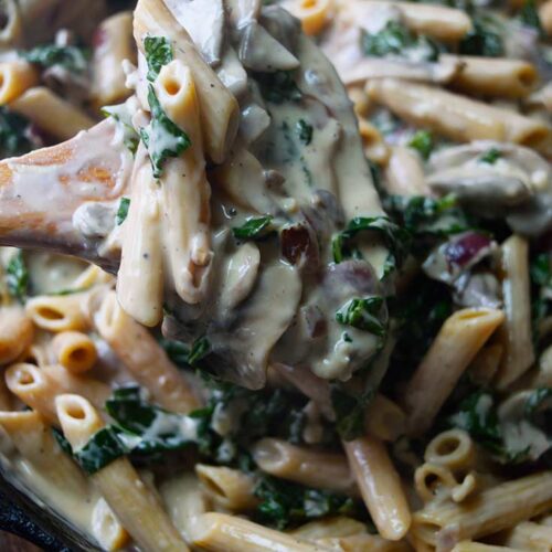 A wooden spoon lifts some Creamy Kale And Mushroom Pasta out of a skillet.