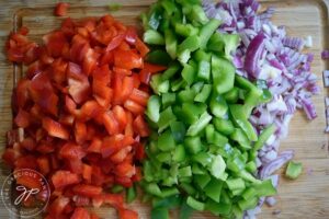Chopped red and green peppers, as well as chopped, red onion laying on a cutting board.