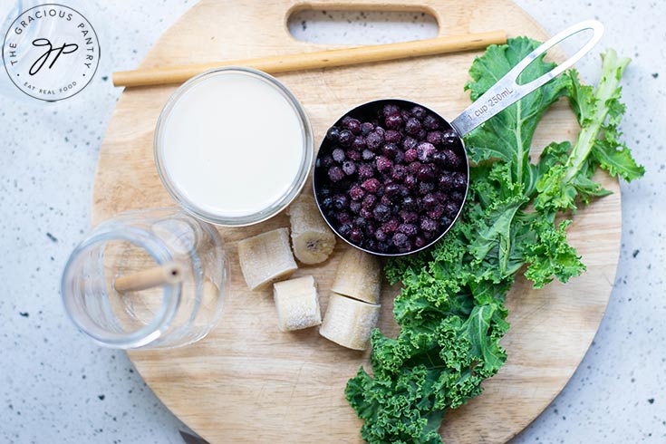 Kale Blueberry Smoothie Recipe ingredients are gathered on a round, wooden, cutting board.
