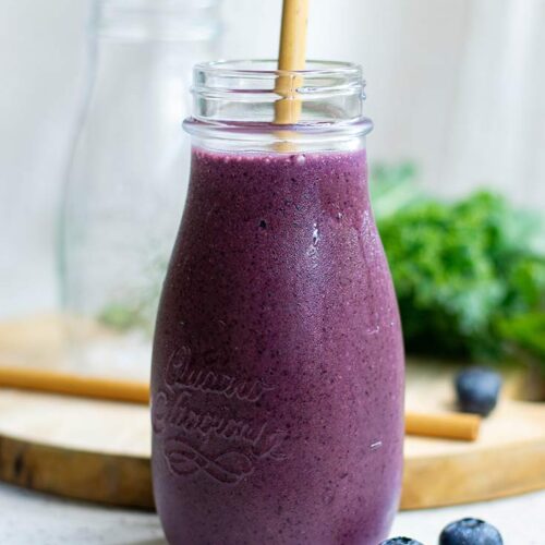 A single glass milk jug sits filled with Kale Blueberry Smoothie. A straw sits in the jar and smoothie as well.