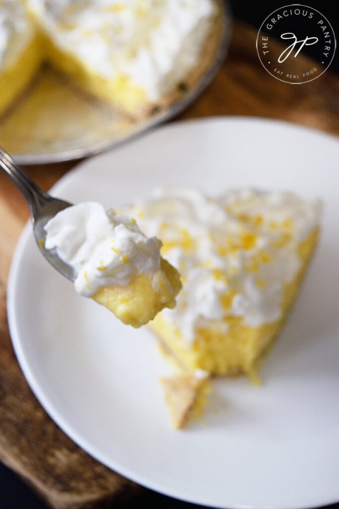 A fork lifts a bit of lemon cream pie off a slice sitting on a white plate.