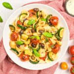 A white bowl filled with Orecchiette Pasta Salad sitting on a red dishtowel, surrounded by fresh basil leaves and cherry tomatoes.