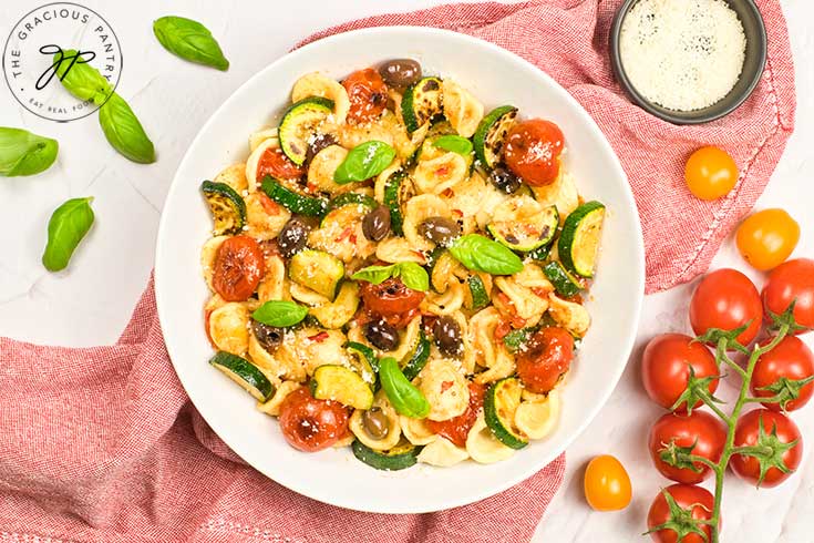 A white bowl filled with Orecchiette Pasta Salad sitting on a red dishtowel, surrounded by fresh basil leaves and cherry tomatoes.