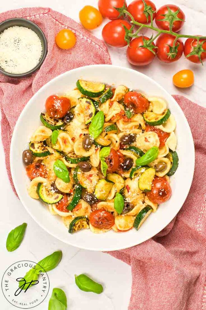 An overhead view of a white bowl filled with Orecchiette Pasta Salad, sitting on a red dishtowel, surrounded by fresh tomatoes and fresh basil leaves.