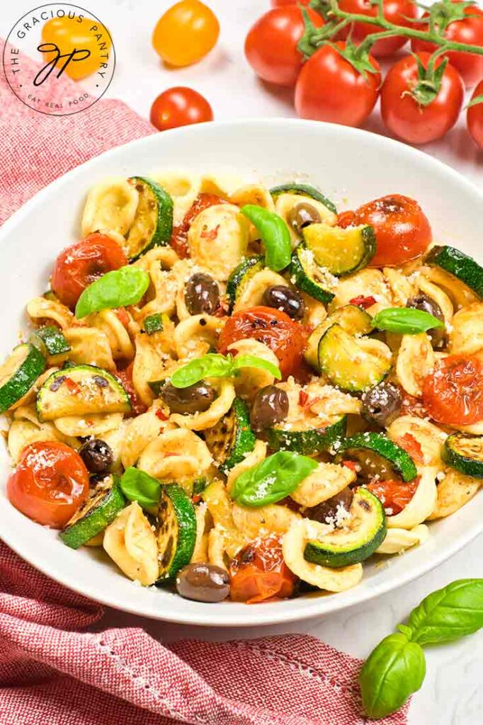 An overhead view of a white bowl filled with Orecchiette Pasta Salad sitting on a rustic red dishtowel.