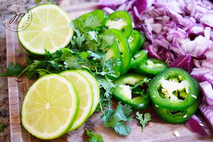 Slow Cooker Cilantro Lime Chicken garnishes sitting on a wood cutting board. Lime slices, chopped cilantro, jalapeno pepper slices and chopped, red onion.