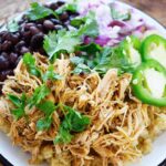 A white plate holds Slow Cooker Cilantro Lime Chicken served over quinoa with black beans, fresh cilantro, jalapeno slices and chopped, red onion.