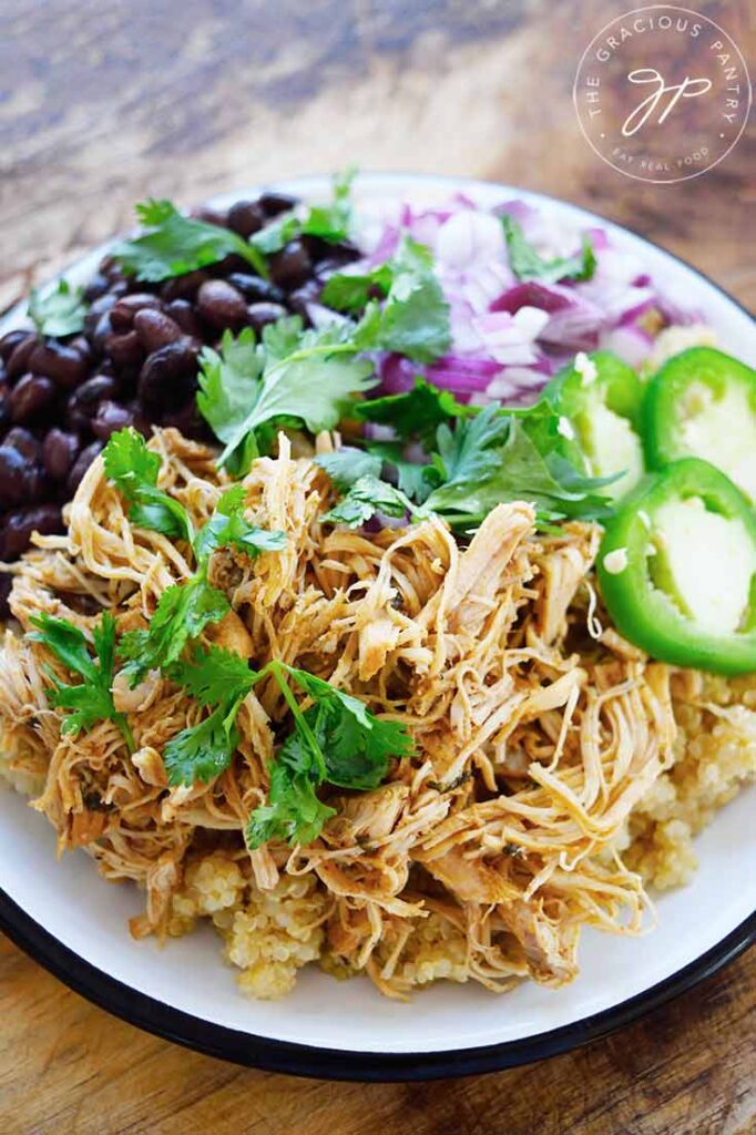 A white plate holds Slow Cooker Cilantro Lime Chicken served over quinoa with black beans, fresh cilantro, jalapeno slices and chopped, red onion.