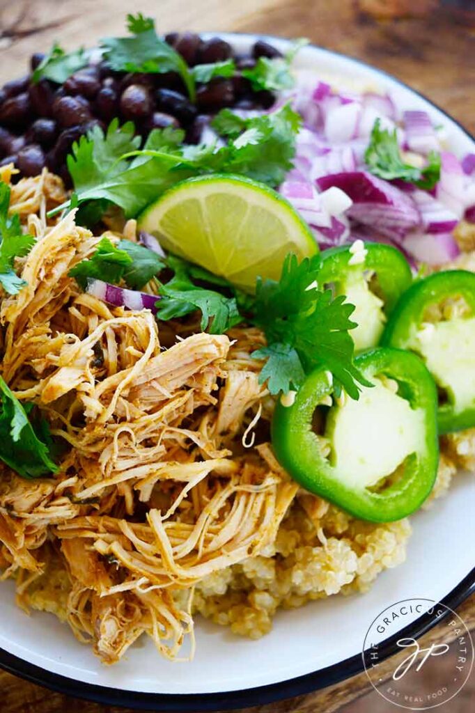 A closeup of a plate of Slow Cooker Cilantro Lime Chicken served over quinoa and garnished with cilantro, lime, jalapeno, red onion and black beans.