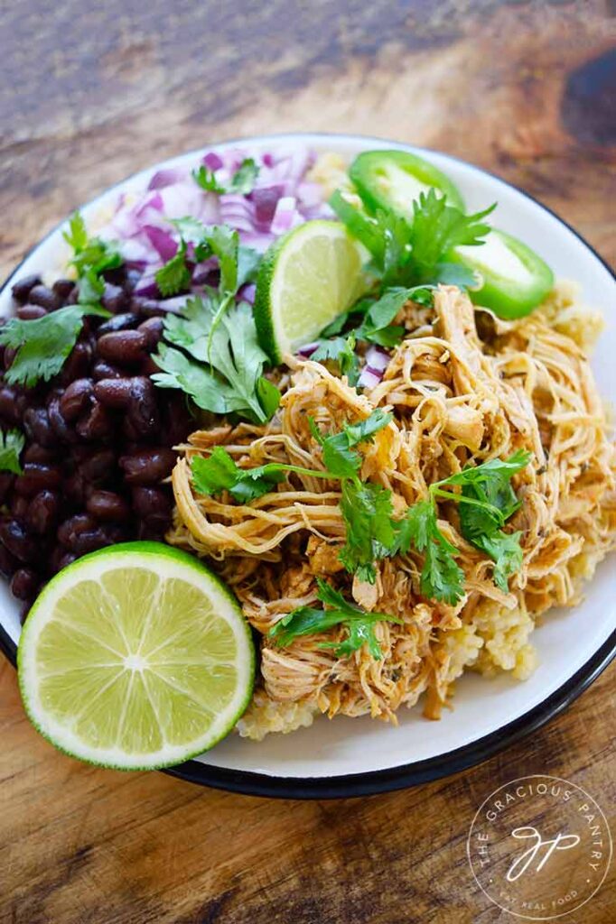 A white plate on a wood surface holds Slow Cooker Cilantro Lime Chicken, quinoa, lime, black beans, jalapeno pepper slices, red onion and fresh cilantro.
