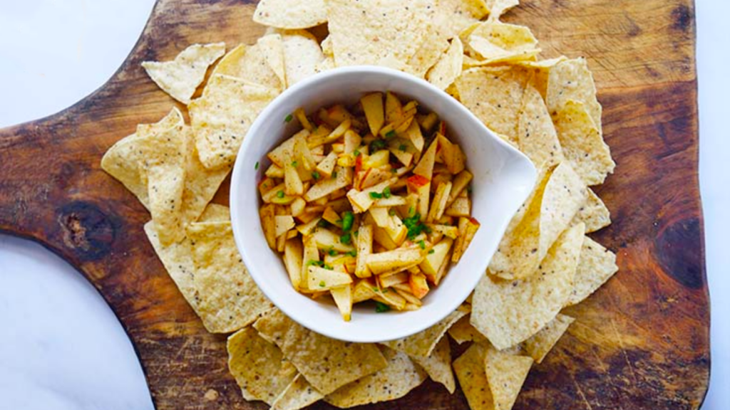 An overhead view of a white bowl filled with apple salsa and surrounded by corn chips on a wood cutting board.