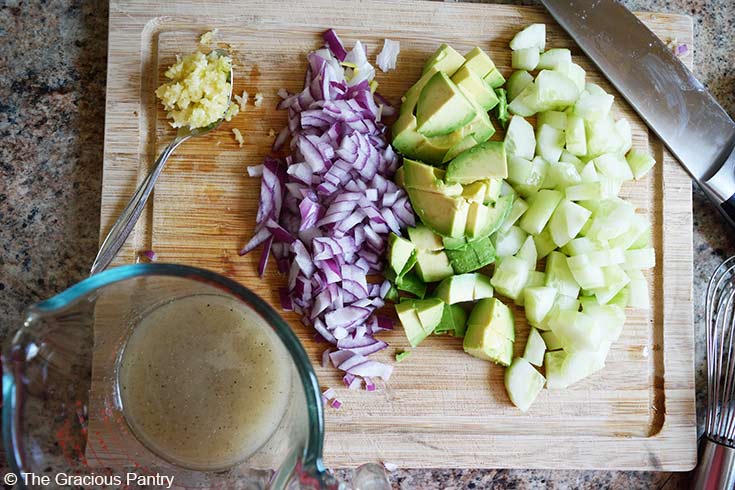 An overhead view of chopped cucumbers, avocado, red onion and press garlic on a cutting board.