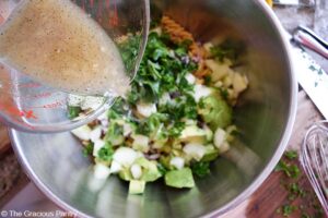 Pouring dressing over Avocado Pasta Salad in a large, metal mixing bowl.