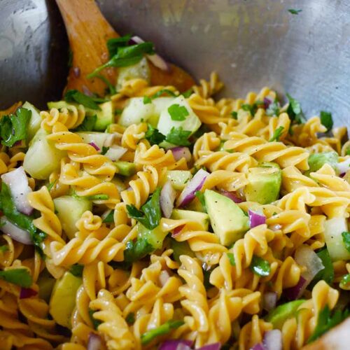 A closeup side view of a large mixing bowl filled with Avocado Pasta Salad. A wooden spoon rests in the bowl.