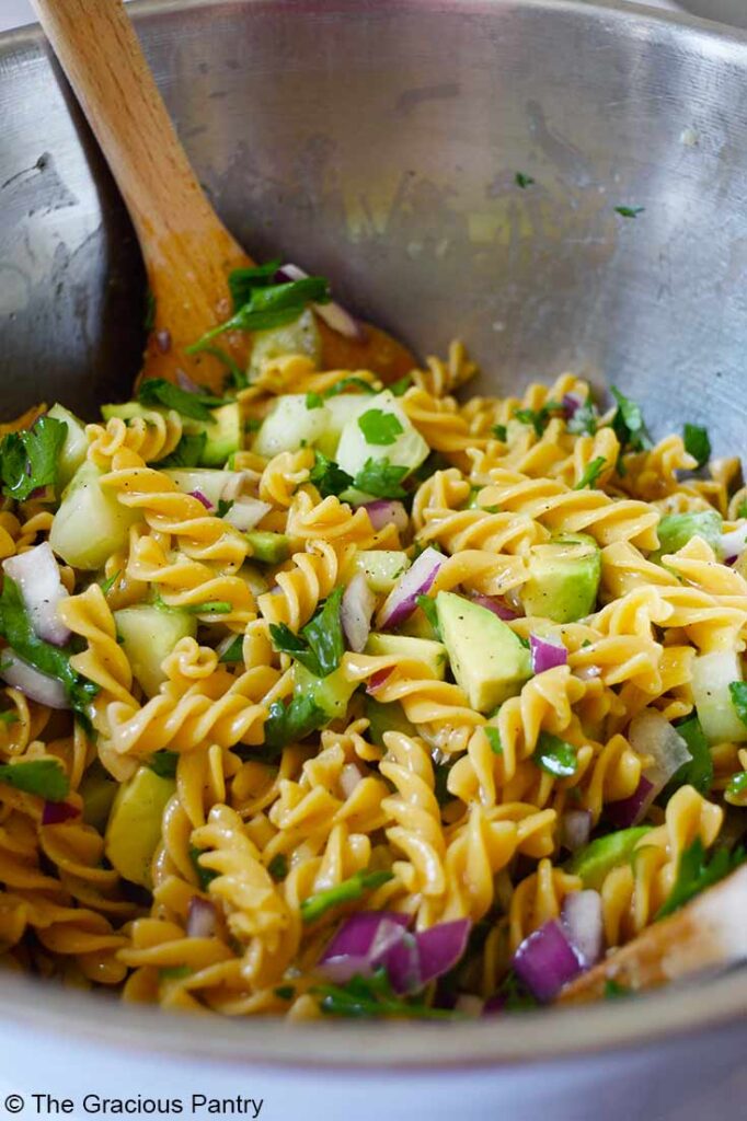 A closeup side view of a large mixing bowl filled with Avocado Pasta Salad. A wooden spoon rests in the bowl.