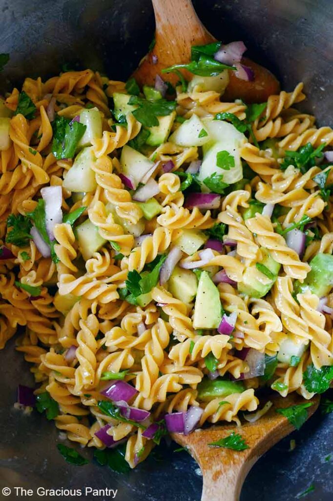 Two wood salad spoons rest in a bowl of Avocado Pasta Salad.