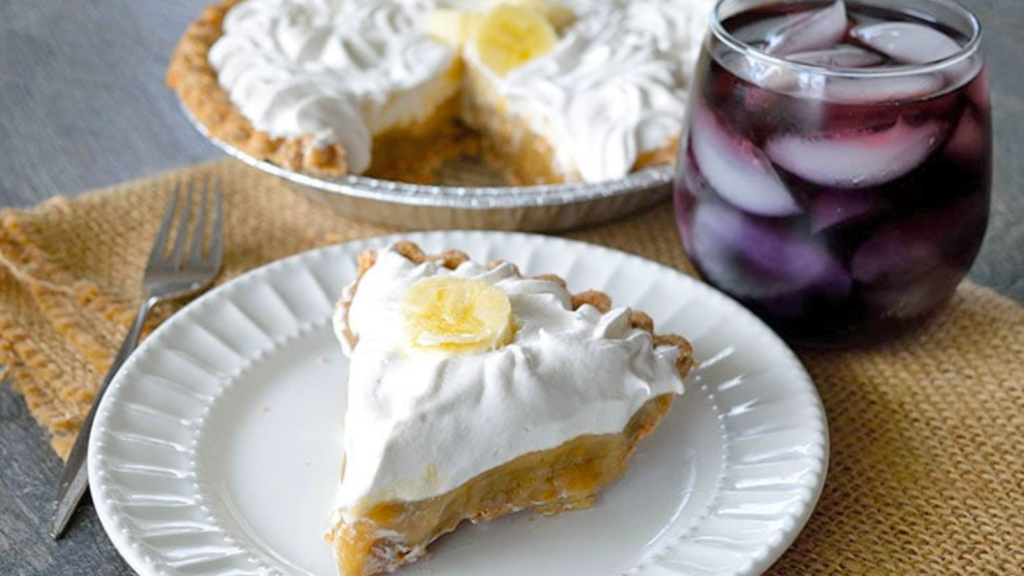 A slice of banana cream pie on a white plate. A drink sits to the right and the rest of the pie sits at the back.