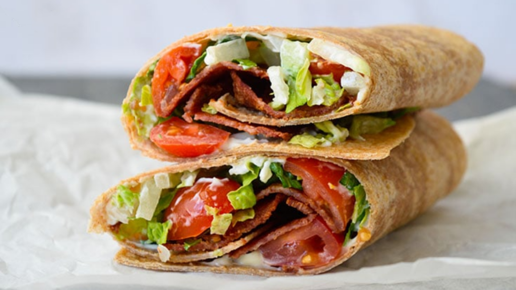 Two BLT wrap halves sit stacked on top of each other on a piece of white parchment paper.
