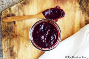 The finished Blueberry BBQ Sauce poured into an open canning jar and sitting on a wood surface next to a wood spoon covered in sauce.