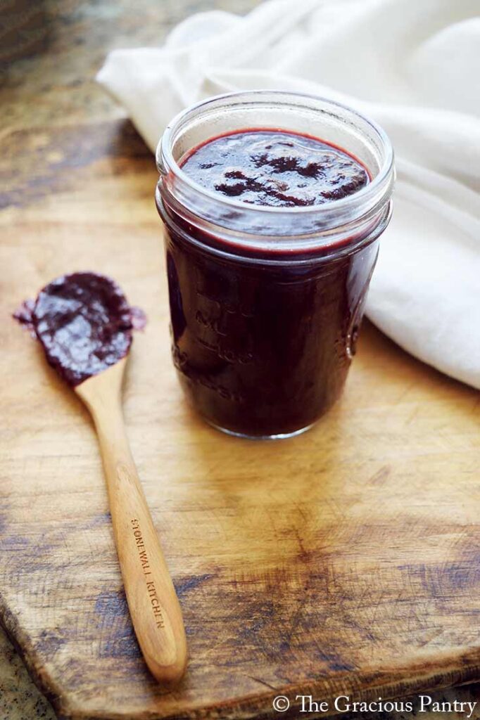 An open canning jar filled with Blueberry BBQ Sauce sits on a cutting board next to a wooden spoon covered in the sauce.