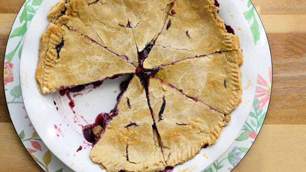 An overhead view of a whole blueberry pie, sliced with one slice missing.