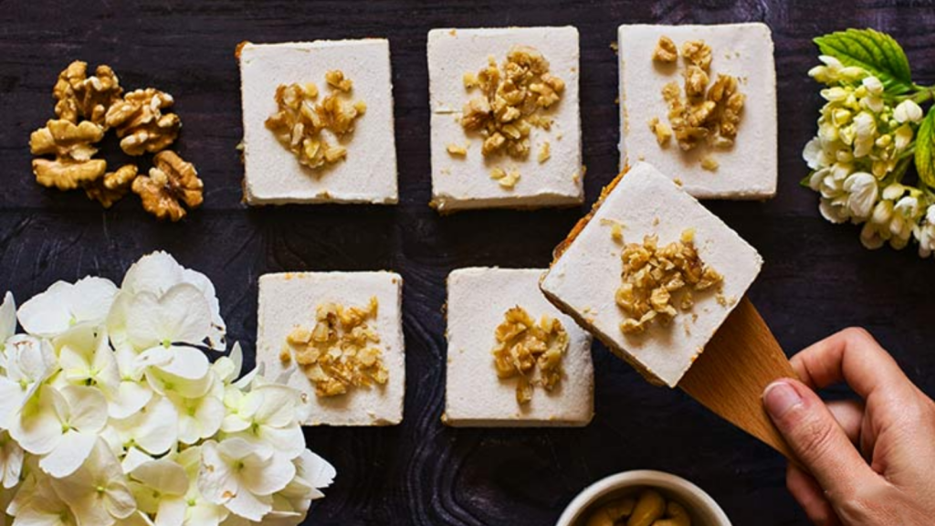 A hand uses a spatula to lift up a piece of carrot cake from a row of square pieces.