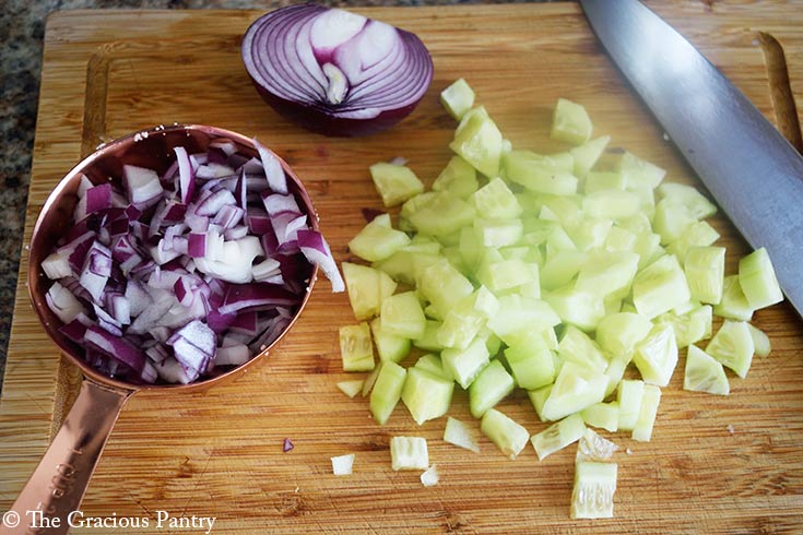 Chopped cucumbers laying on a wood cutting board next to a measure cup filled with chopped red onion.