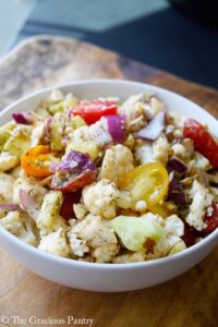 Cauliflower salad in a white bowl. Halved grape tomatoes are seen in both red and yellow colors along with purple onion.