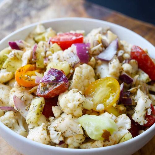 Cauliflower salad in a white bowl. Halved grape tomatoes are seen in both red and yellow colors along with purple onion.