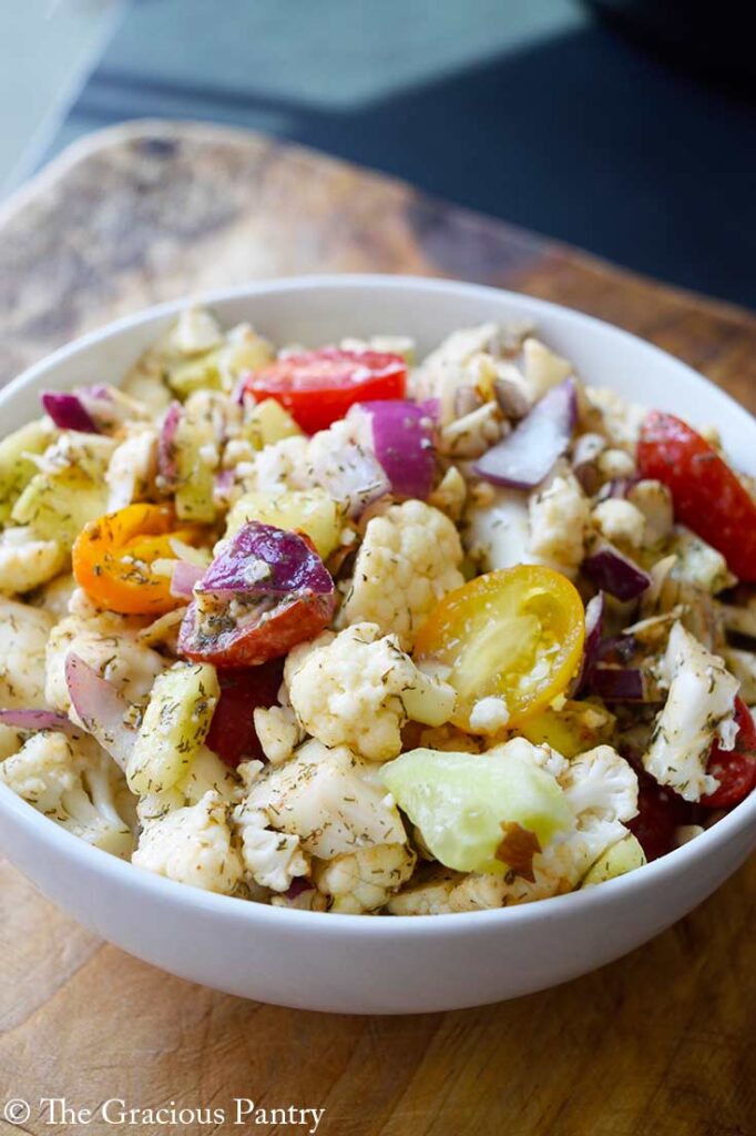 Cauliflower salad in a white bowl. Halved grape tomatoes are seen in both red and yellow colors along with purple onion.