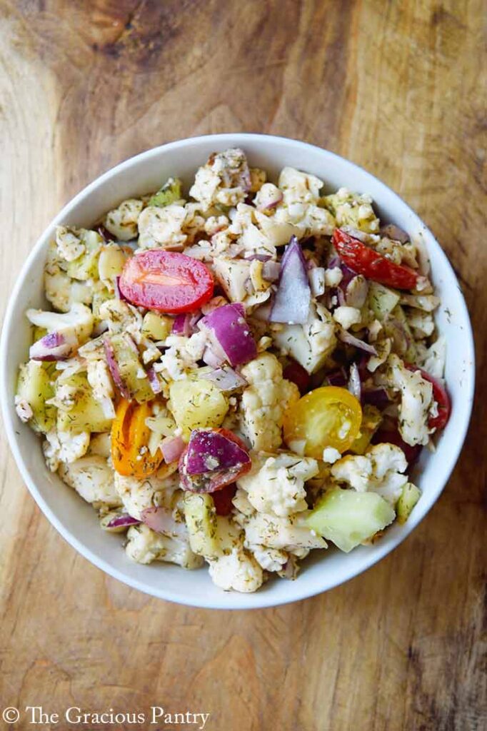 An overhead view looking down into a white bowl on a wood surface, filled with cauliflower salad.