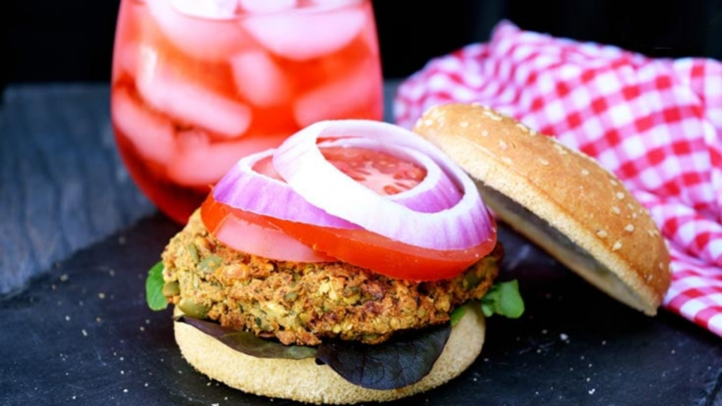 A chickpea burger sits with it's top bun off to the side showing off onion and tomato slices over the chickpea burger patty.