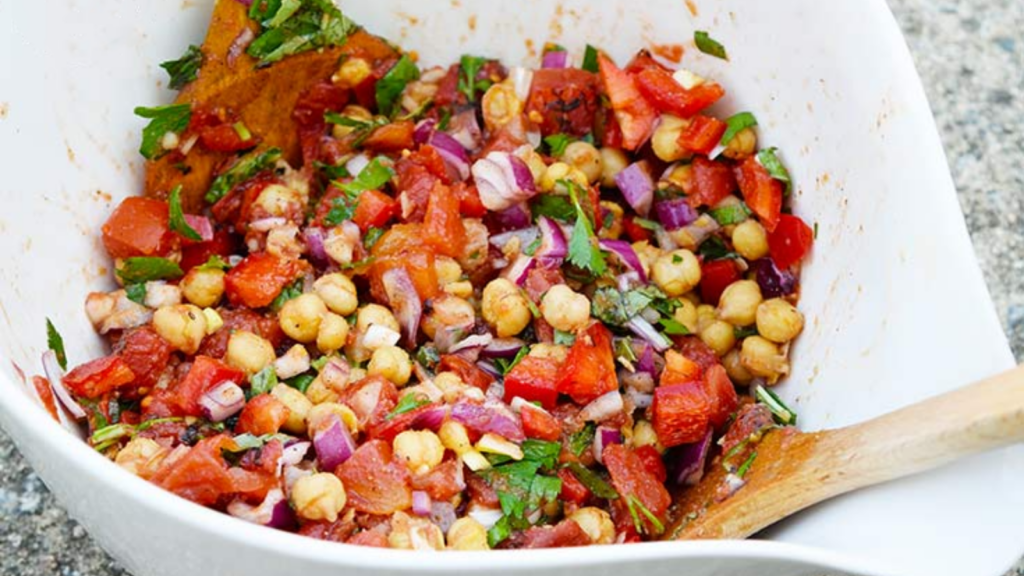 A white ceramic mixing bowl holds some chickpea salad. A wood spoon rests inside the bowl.
