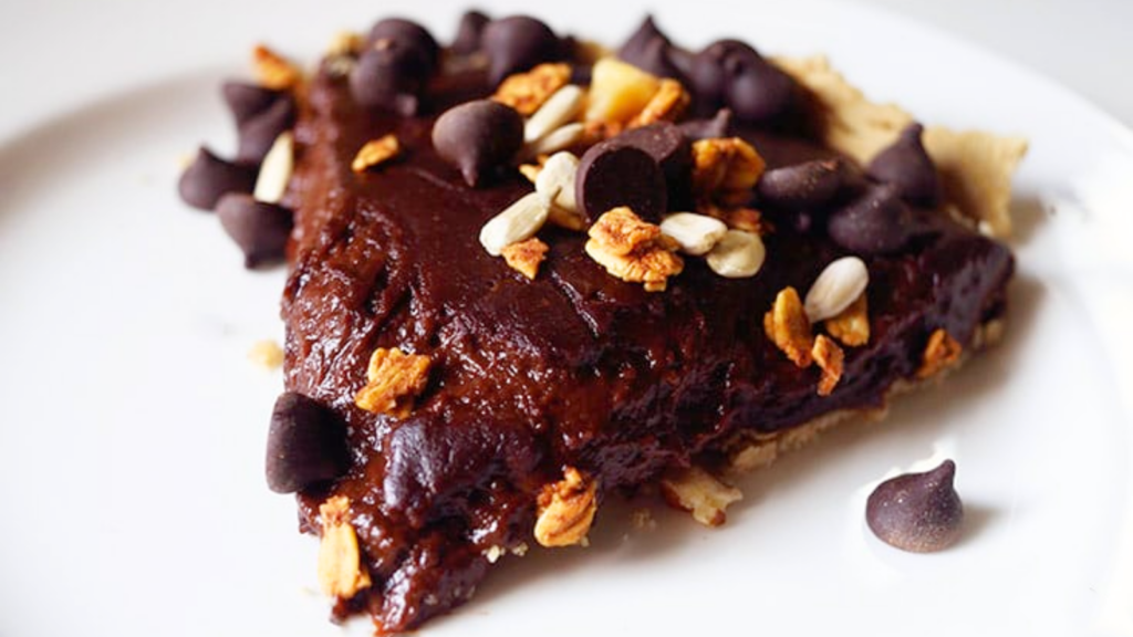 A slice of chocolate pie sits on a white plate, topped with chocolate chips and walnut pieces.