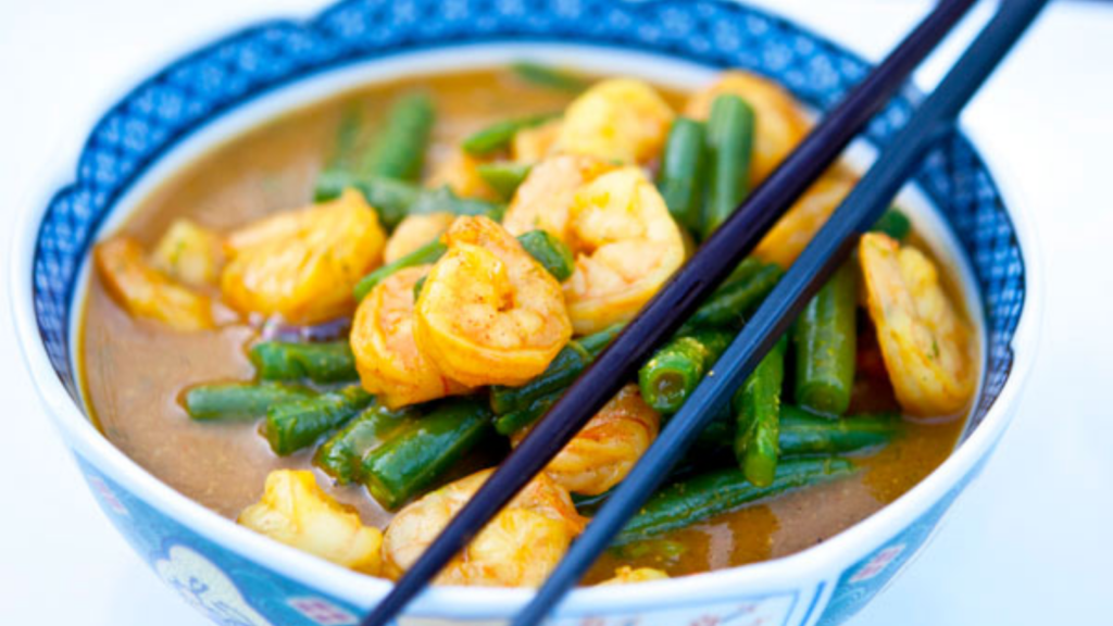A blue bowl holds a serving of coconut curry shrimp with green beans. Black chopsticks lay across the bowl.