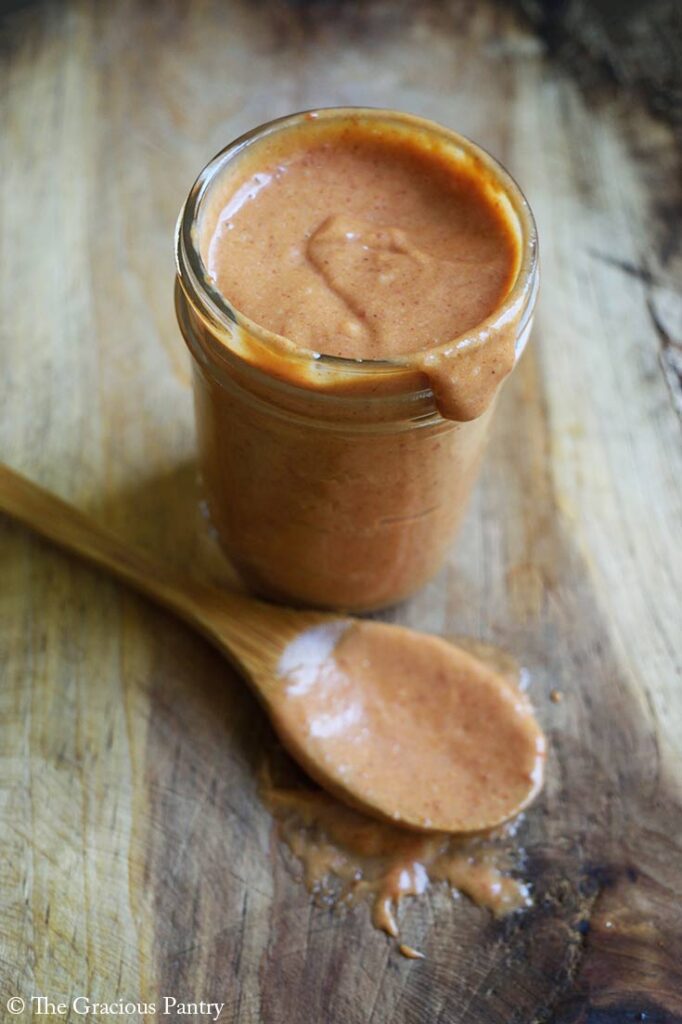 A canning jar sits open and filled with Copycat Chick-Fil-A Honey Roasted Barbecue Sauce. A sauce-covered wooden spoon lays next to it on a wooden surface.