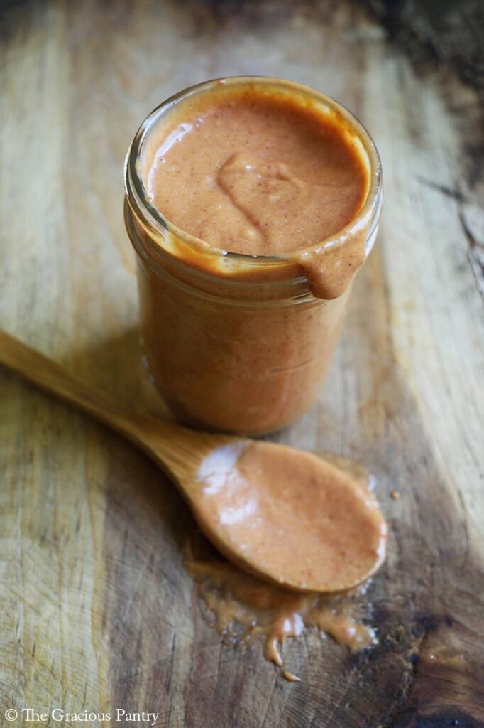 A canning jar sits open and filled with Copycat Chick Fil A Honey BBQ Sauce. A sauce-covered wooden spoon lays next to it on a wooden surface.